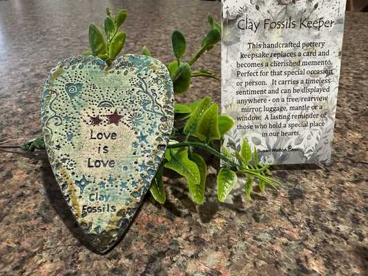 Heart-shaped clay fossil with 'Love is Love' text on a marble surface with a card explaining the product.